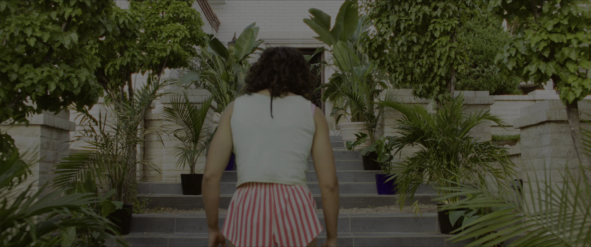 a woman with dark curly hair walks up a set of stairs. she wears a white tank top and pink and white striped shorts